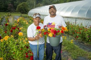 Two farmers stand in a field of cultivated flowers. There is a greenhouse in the background and they are both holding a bouquet of red and yellow flowers.